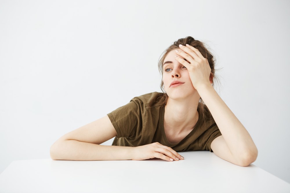 Bored tired dull young girl student with bun sitting at table over white background.
